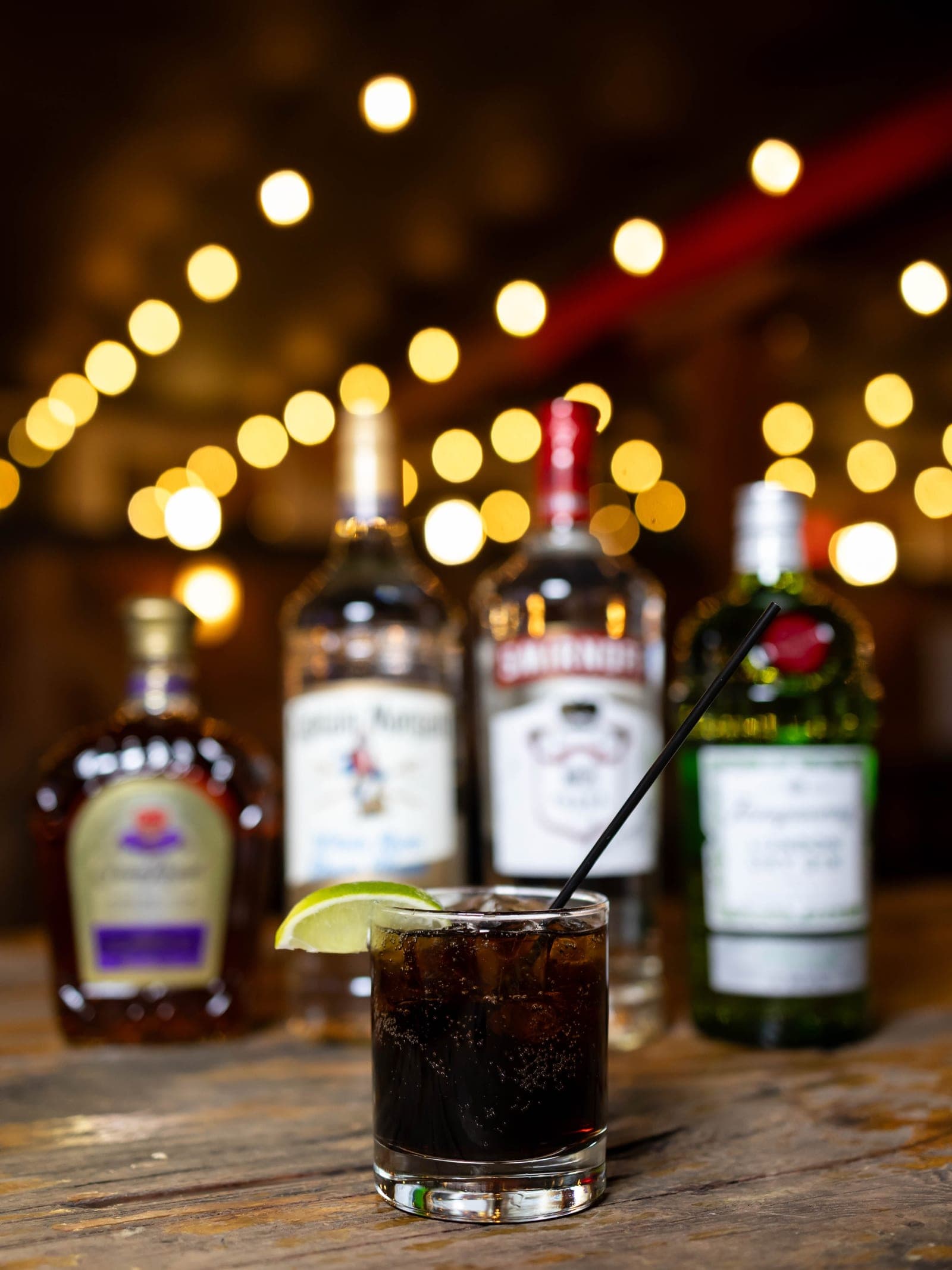 Craft cocktail poured over ice with a backbar of spirits and warm string lights