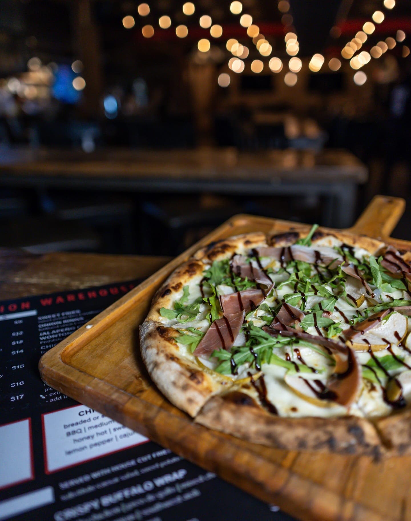 Wood-fired pear and prosciutto pizza on a wooden peel under warm string lights