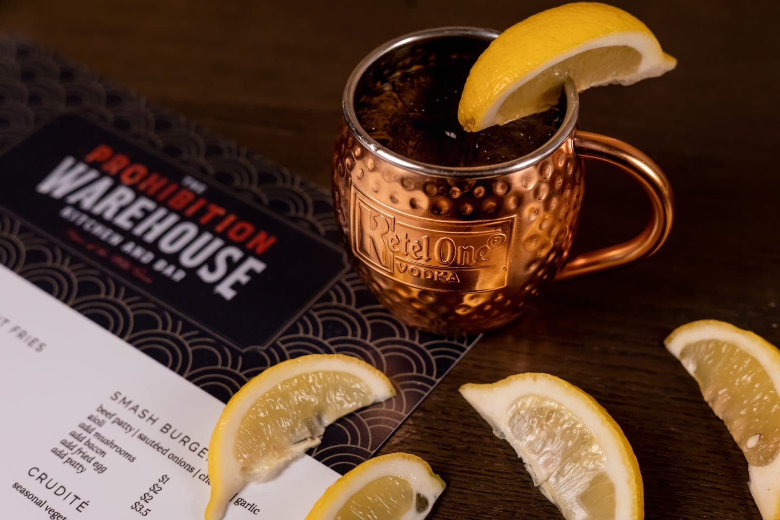 Copper mule mug with lemon beside the Prohibition Warehouse menu on dark wood